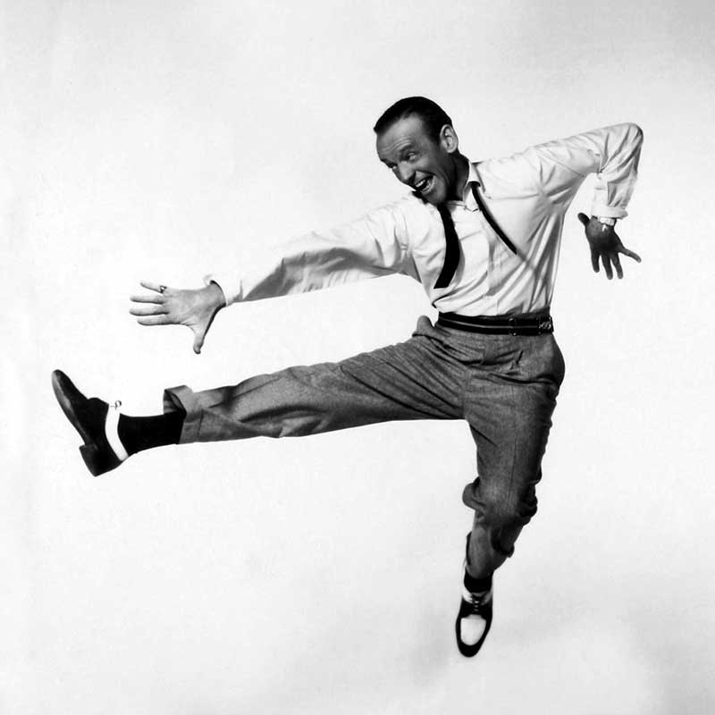 The dancer Fred Astaire is performing a lively jump against a white background while dancing. He's wearing a white shirt, an untied tie, and gray pants.